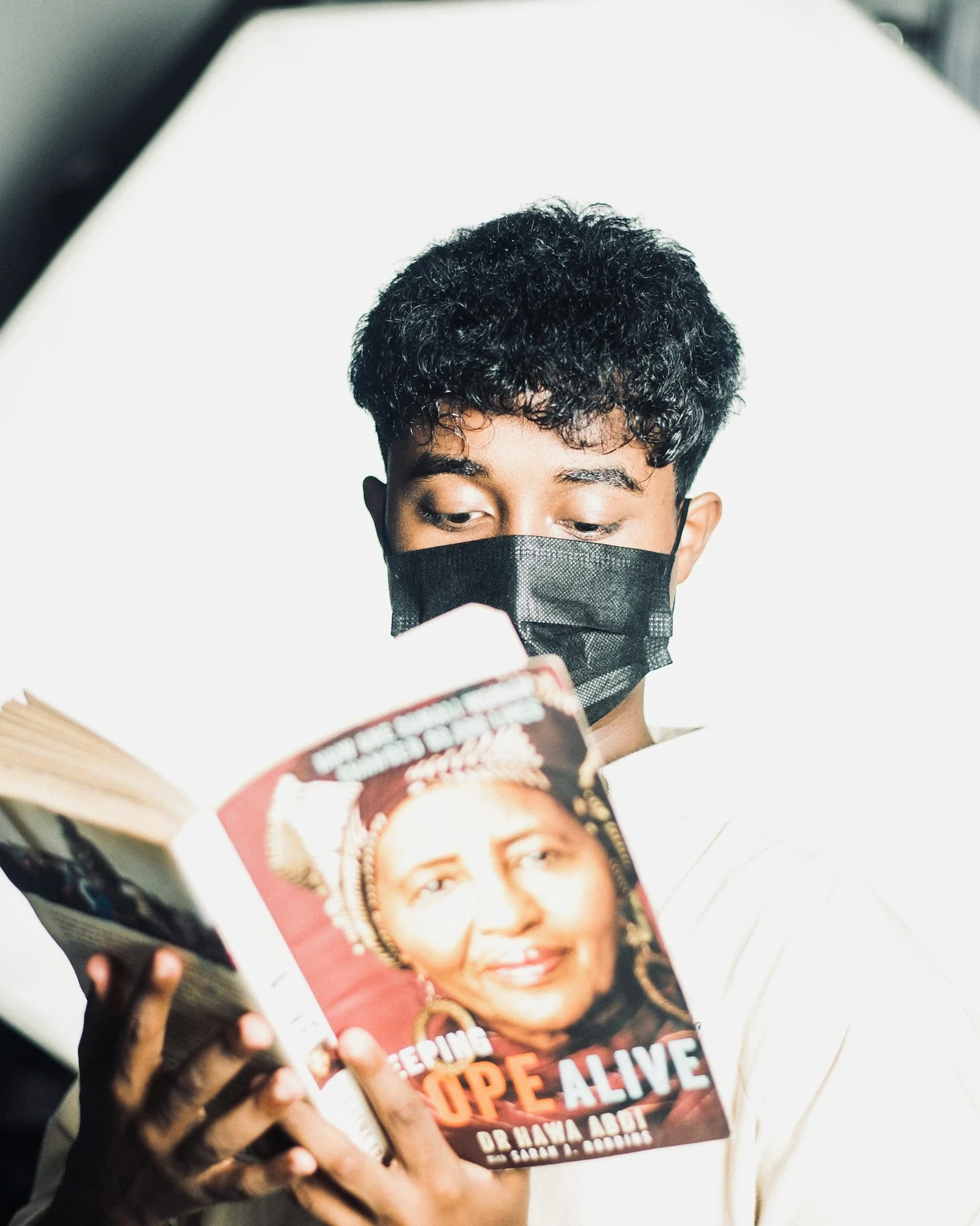 A person in a black mask holding a book in strong sunlight.