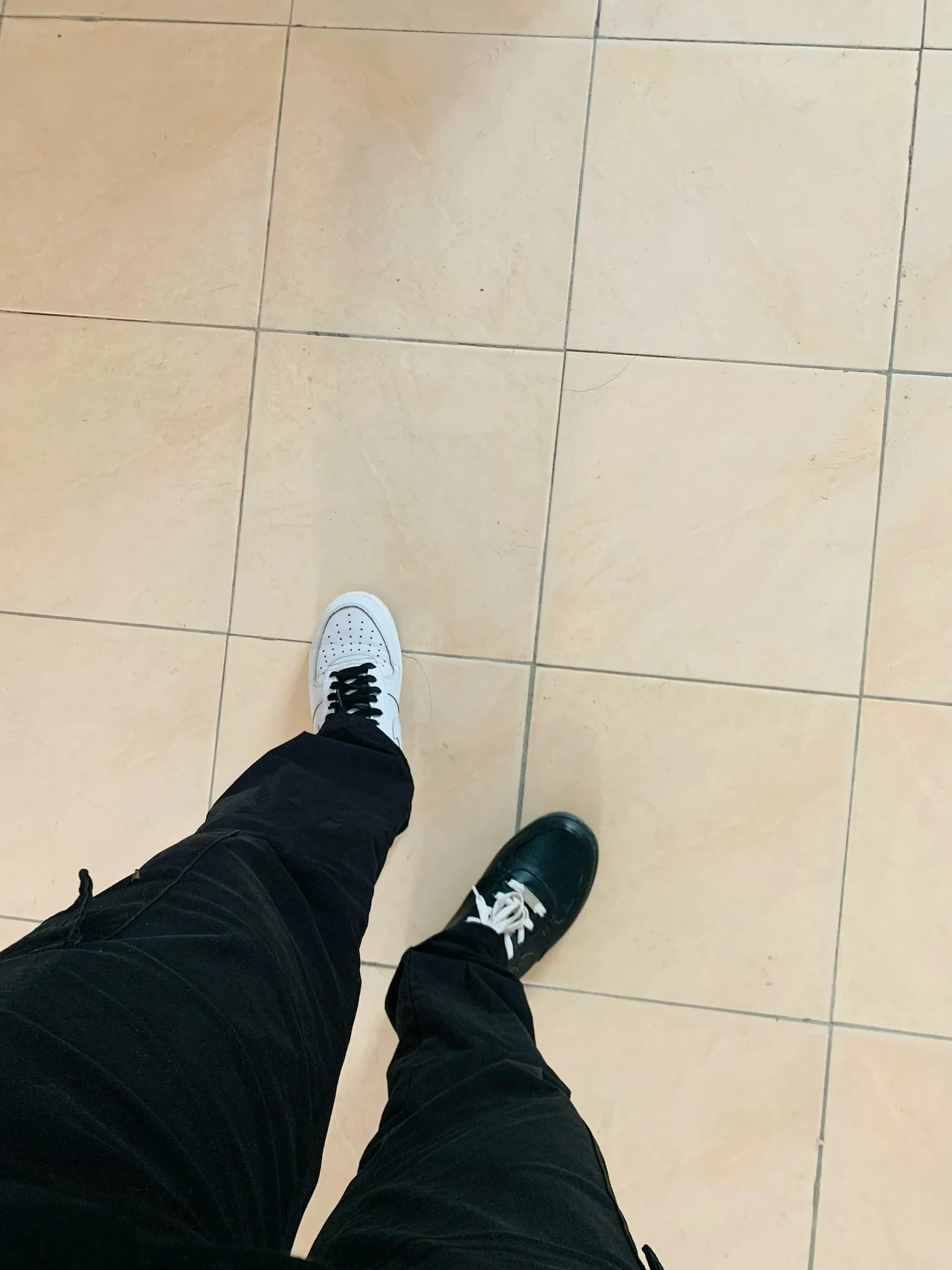 A downward view of black trousers and shoes on pale floor tiles.